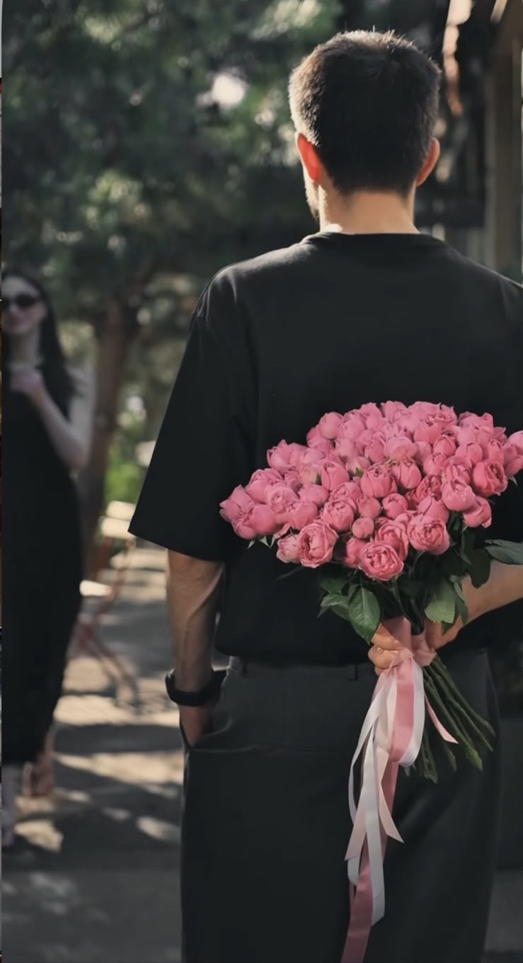 A man holding a large pink rose bouquet behind his back as a woman approaches